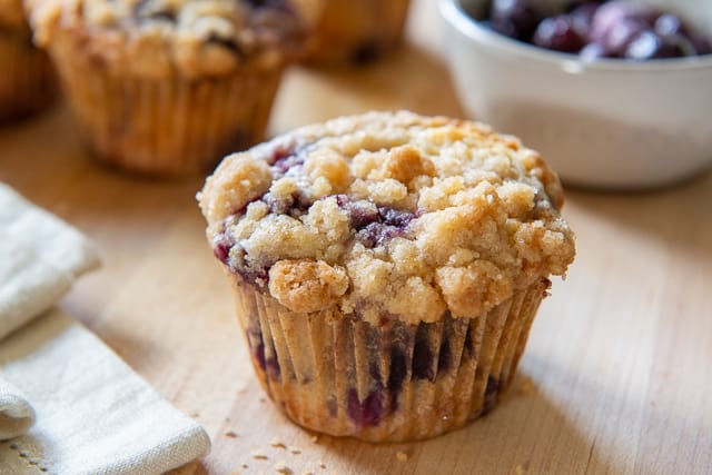 A close up of a blueberry muffin with crunchy golden topping and bowl of blueberries on side