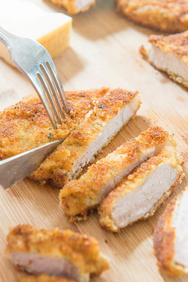 Parmesan Crusted Pork Chops - Served On a Wooden Platter with a Fork and Knife Slicing Into Pieces
