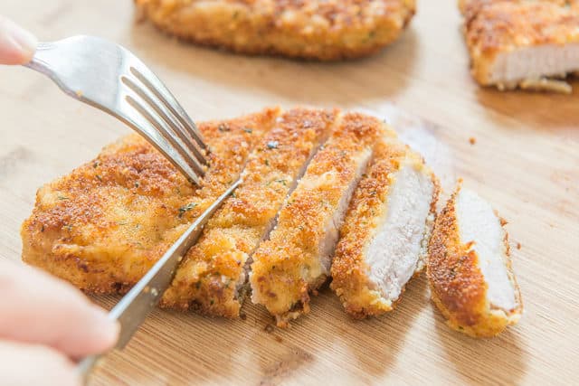 Parmesan Pork Chops - Served On a Wooden Platter with a Fork and Knife Slicing Into Pieces