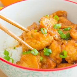 Chopsticks Holding a Piece of Orange Chicken Garnished with Scallions in a Bowl with White Rice
