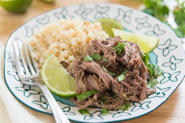 A Cup Of Shredded Indian Spiced Beef With Cauliflower Rice, Lime Wedges, Cilantro, And a Fork