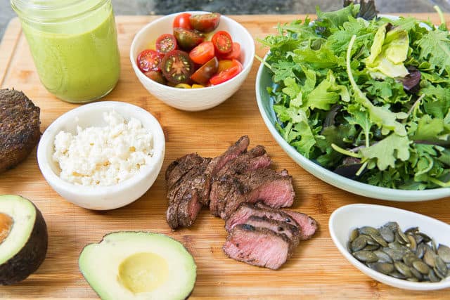 Steak Salad Ingredients on Wooden Board Including Greens, Avocado, Cheese, Tomatoes, and Dressing