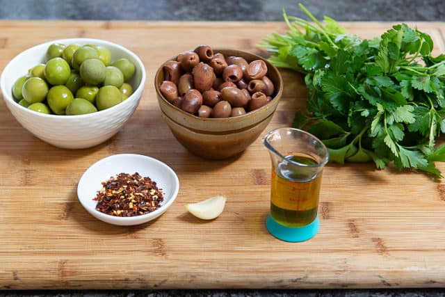Olive Tapenade Recipe Ingredients on a Cutting Board with Olive Oil, Garlic, Pepper Flakes, Olives, and Parsley