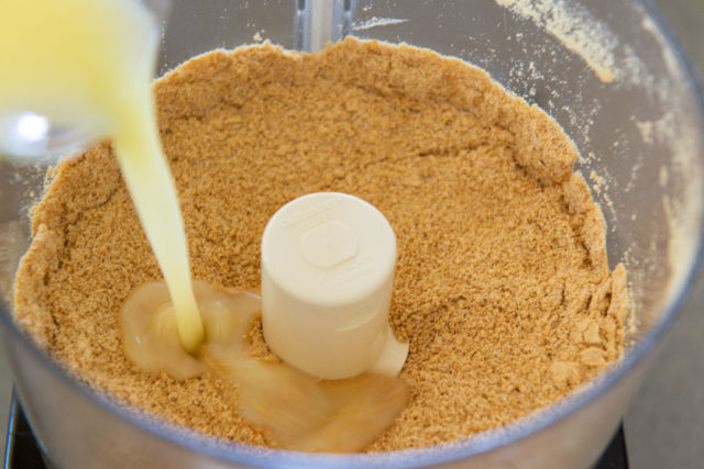 Graham Cracker Crumbs in Food Processor Bowl with Melted Butter Pouring In