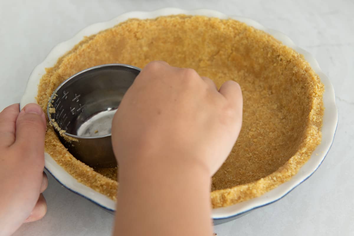 Using a measuring cup to firmly compact the cookie crumbs up the sides of the pie plate