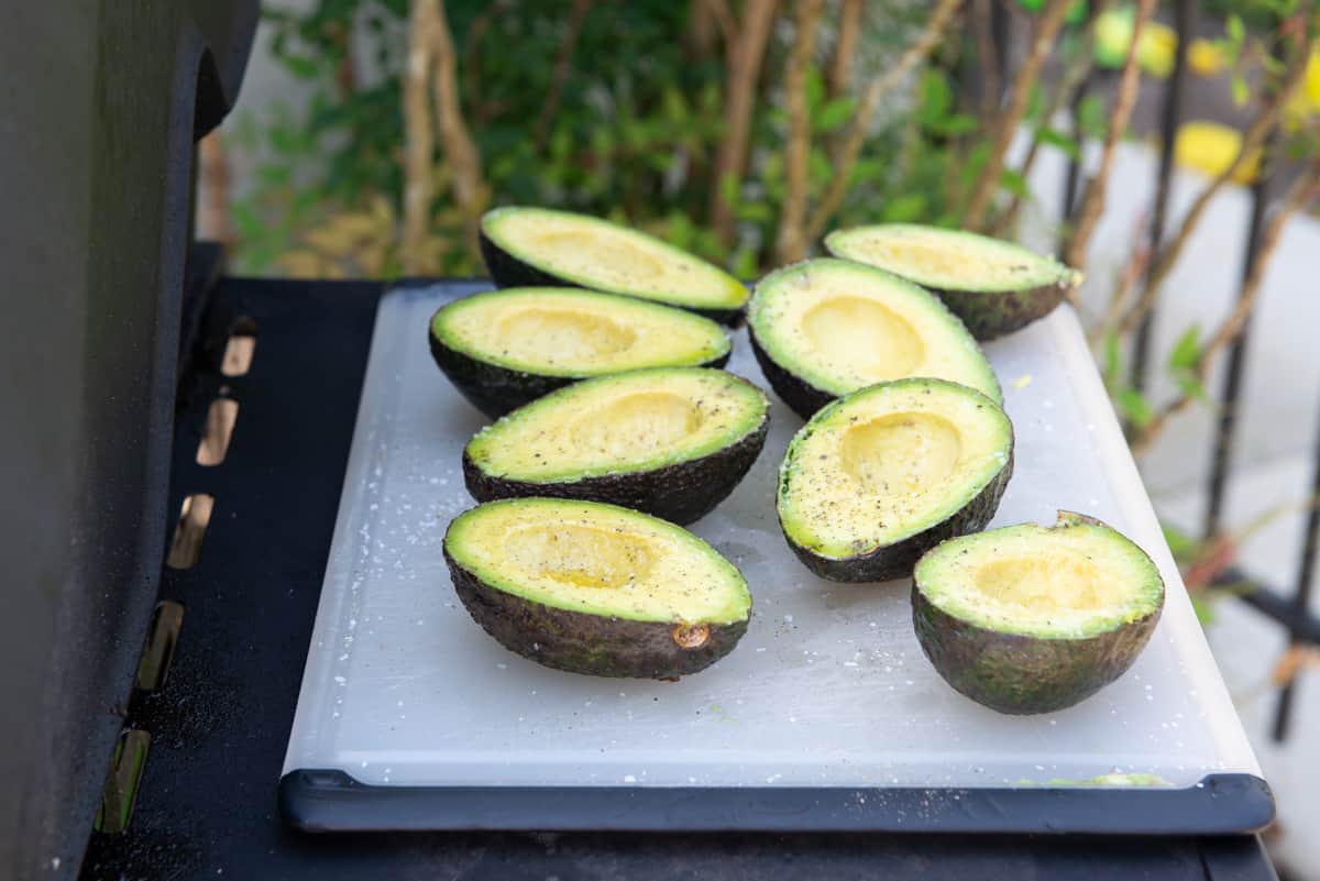 Cut Avocado Halves On Cutting Board On Grill Shelf with Salt, Pepper, and Olive Oil On Top