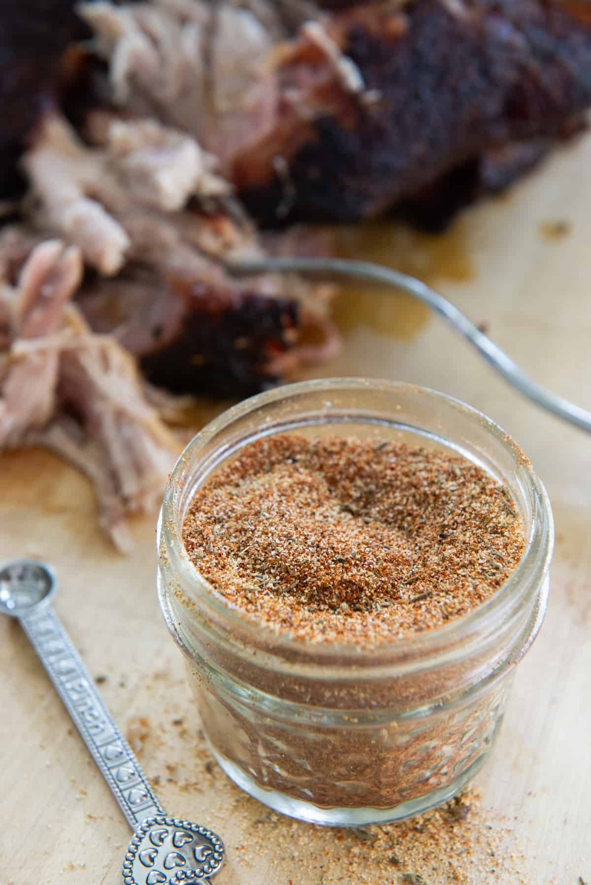 A small glass jar of pulled pork rub with pulled pork butt in background
