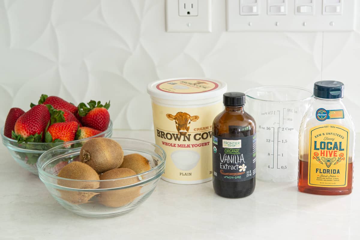 A bowl of strawberries, kiwis, yogurt, vanilla extract, ice, and honey on a countertop