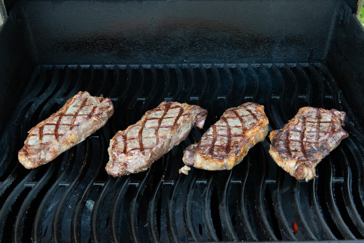 4 NY Strip Steaks On Grill Grates Of Gas Grill With Crosshatch Marks On Top