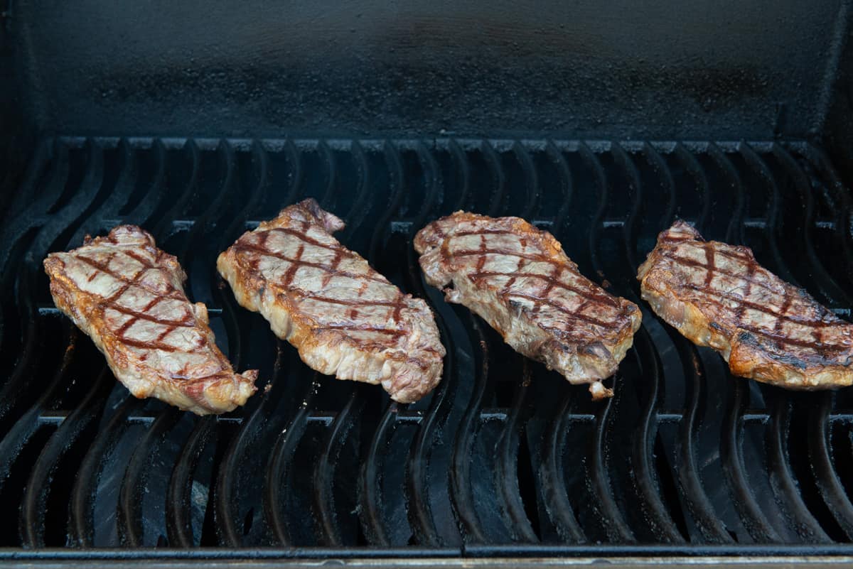 4 NY Strip Steaks On Grill Grates Of Gas Grill With Crosshatch Marks On Top Going Other Way