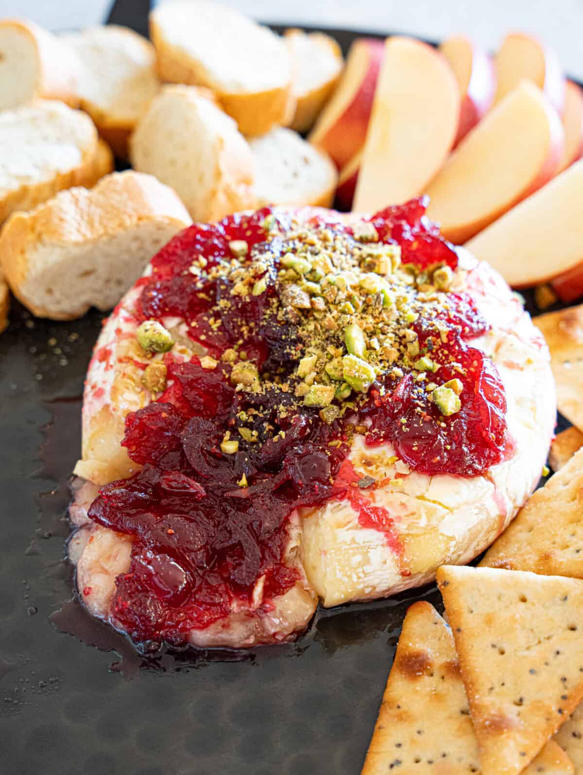 Gooey Brie cheese topped with cranberry sauce, pistachios and surrounded by apple slices, crackers, and bread