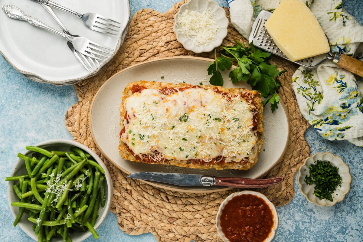 Parmesan chicken meatloaf on an oval platter in the middle of a table setting with a bowl of steamed green beans nearby.