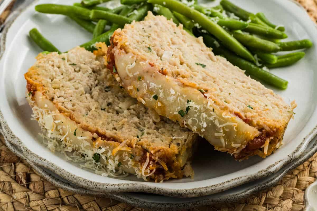 Two slices of parmesan chicken meatloaf arranged next to steamed green beans on a plate.