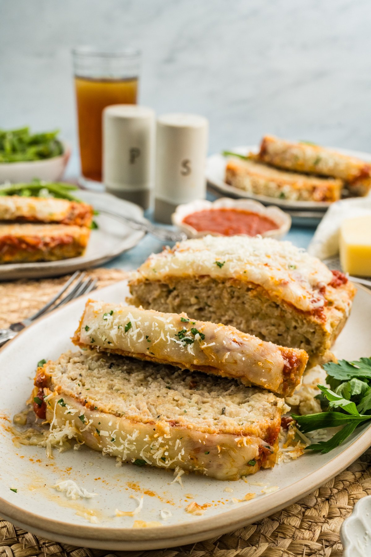 Platter of sliced parmesan chicken meatloaf on a table with plates of meatloaf and salt and pepper shakers in the background.